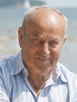 L&auml;chelnder &auml;lterer Mann mit Hemd im Streifenmuster vor unscharfem Wasserhintergrund.