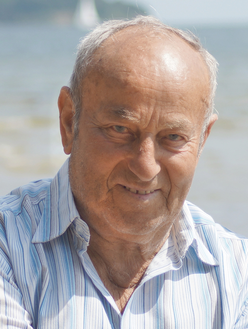 L&auml;chelnder &auml;lterer Mann mit Hemd im Streifenmuster vor unscharfem Wasserhintergrund.