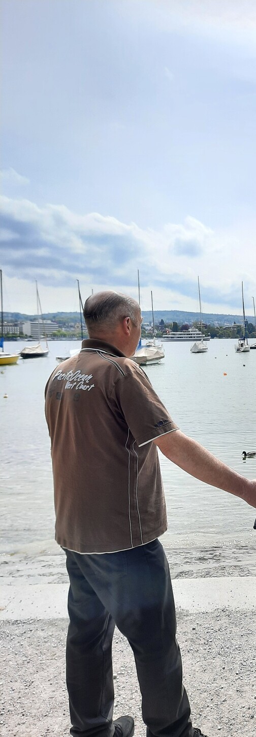 Mann steht am Ufer eines Gew&auml;ssers und blickt auf Boote, w&auml;hrend eine Ente im Wasser schwimmt.