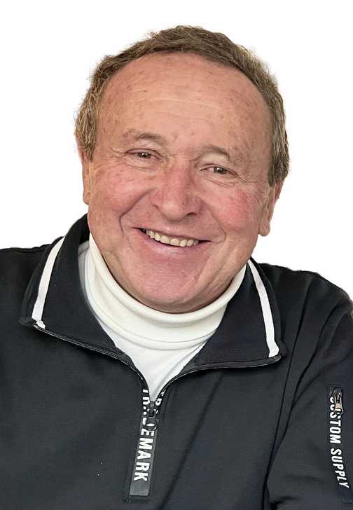 L&auml;chelnder &auml;lterer Mann in schwarzer Zip-Jacke mit wei&szlig;em Rollkragenpullover vor einem wei&szlig;en Hintergrund.
