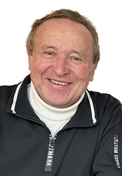 L&auml;chelnder &auml;lterer Mann in schwarzer Zip-Jacke mit wei&szlig;em Rollkragenpullover vor einem wei&szlig;en Hintergrund.