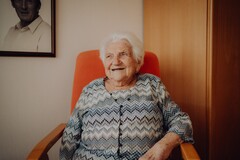 Ältere Frau mit weißem Haar sitzt lächelnd in einem orangefarbenen Sessel, im Hintergrund ein schwarzes undweißes Foto.