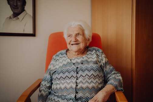 &Auml;ltere Frau mit wei&szlig;em Haar sitzt l&auml;chelnd in einem orangefarbenen Sessel, im Hintergrund ein schwarzes undwei&szlig;es Foto.