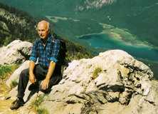 Ein älterer Mann in blauer Karobluse sitzt auf einem Felsen mit Blick auf ein grünes Tal und einen blauen See.