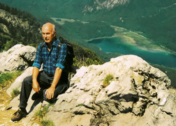 Ein &auml;lterer Mann in blauer Karobluse sitzt auf einem Felsen mit Blick auf ein gr&uuml;nes Tal und einen blauen See.