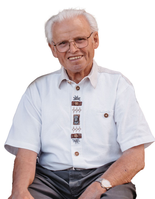 &Auml;lterer Mann mit Brille in wei&szlig;em Hemd mit dekorativen Kn&ouml;pfen sitzt l&auml;chelnd auf einem Stuhl.