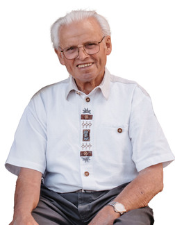 &Auml;lterer Mann mit Brille in wei&szlig;em Hemd mit dekorativen Kn&ouml;pfen sitzt l&auml;chelnd auf einem Stuhl.