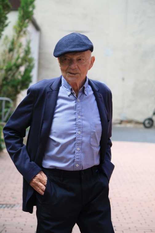 &Auml;lterer Mann mit Schirmm&uuml;tze und blauer Jacke steht entspannt in einer urbanen Umgebung.