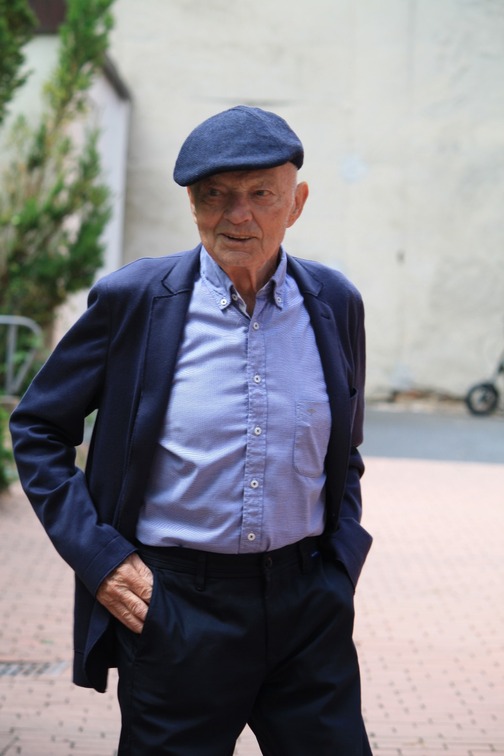 &Auml;lterer Mann mit Schirmm&uuml;tze und blauer Jacke steht entspannt in einer urbanen Umgebung.