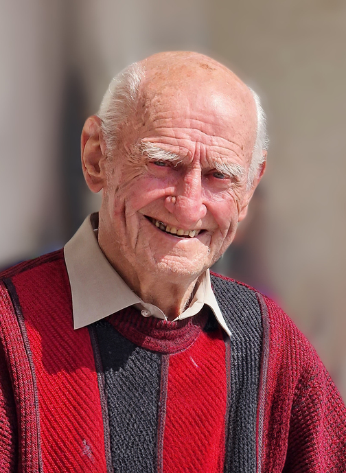 L&auml;chelnder &auml;lterer Mann mit Glatze in rot-schwarzem Pullover vor unscharfem Hintergrund.