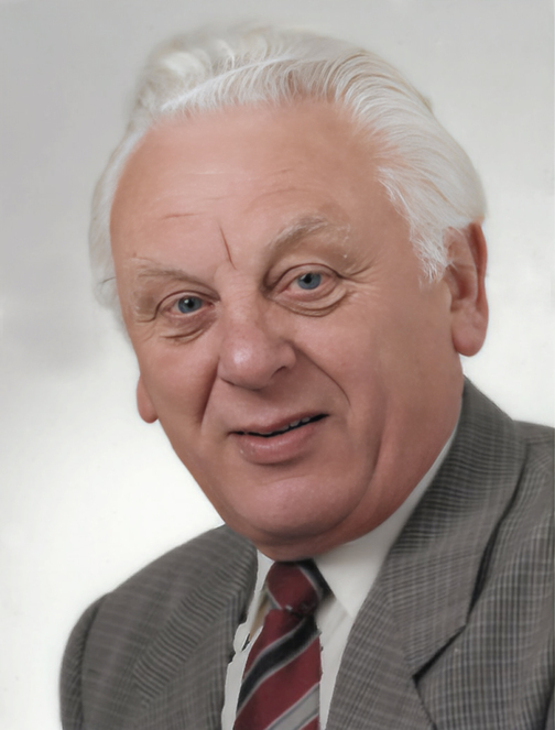 Portr&auml;t eines &auml;lteren Mannes mit grauen Haaren, in einem karierten Anzug und gestreifter Krawatte, vor hellgrauem Hintergrund.