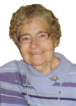 L&auml;chelnde &auml;ltere Frau mit Brille, grau-blonden Locken und lila gestreiftem Pullover, tr&auml;gt eine Perlenkette und Brosche.