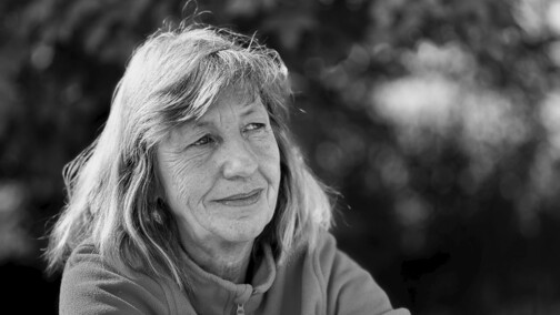 Portr&auml;t einer l&auml;chelnden Frau mit grauen Haaren, sitzend vor unscharfem, gr&uuml;nem Hintergrund.