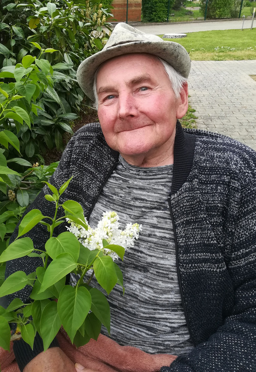 &Auml;lterer Mann mit grauem Hut h&auml;lt einen Zweig mit wei&szlig;en Bl&uuml;ten und gr&uuml;nen Bl&auml;ttern, sitzt neben einer Pflanze.