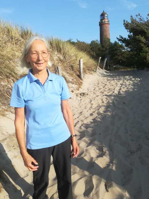 Frau mit langen, grauen Haaren steht am Strandweg, im Hintergrund ein roter Leuchtturm und gr&uuml;ne B&auml;ume.