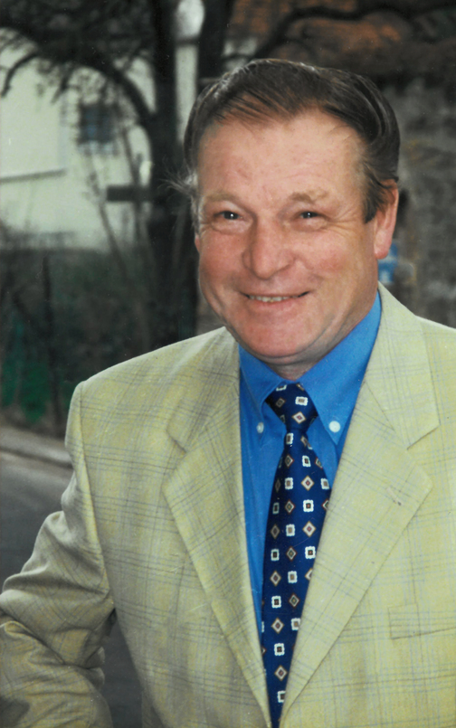 L&auml;chelnder Mann in beigem Blazer mit gemusterter Krawatte und blauem Hemd, vor unscharfem Hintergrund im Freien.