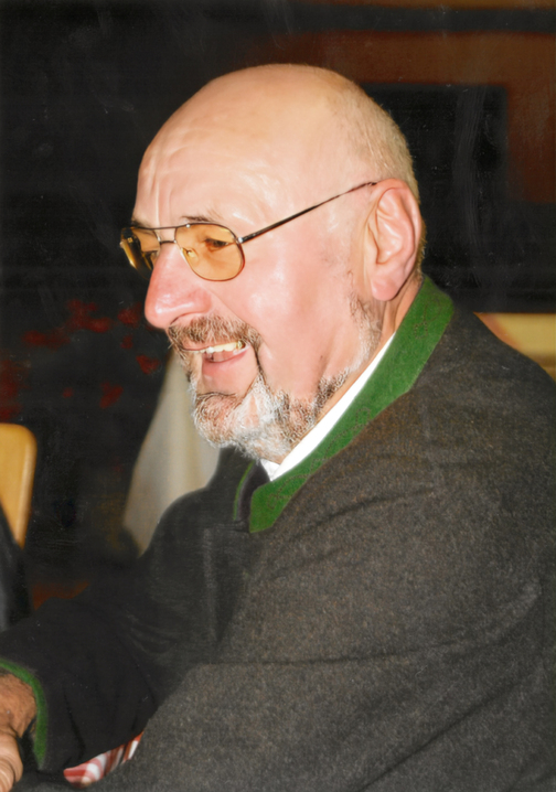 Portr&auml;t eines l&auml;chelnden &auml;lteren Mannes mit Glatze und Bart, tr&auml;gt eine Brille und einen dunklen Pullover mit gr&uuml;nem Kragen.