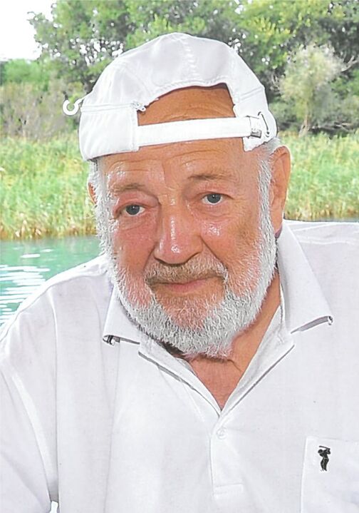 Portr&auml;t eines &auml;lteren Mannes mit wei&szlig;em Kapit&auml;nshut und Bart, am Ufer eines Gew&auml;ssers. Gr&uuml;ne Vegetation im Hintergrund.