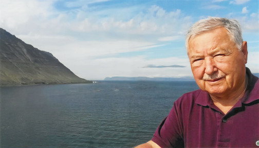 Ein &auml;lterer Mann mit grauem Haar steht am Wasser, im Hintergrund sind Berge und ein klarer Himmel zu sehen.