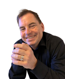 Portr&auml;t eines Mannes mit kurzem Haar, der in einem schwarzen Hemd l&auml;chelt und seine H&auml;nde zusammengelegt hat.