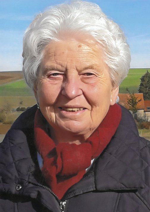 Portrait einer &auml;lteren Frau mit grauen Haaren, die l&auml;chelt und einen roten Schal tr&auml;gt, vor einer l&auml;ndlichen Landschaft.