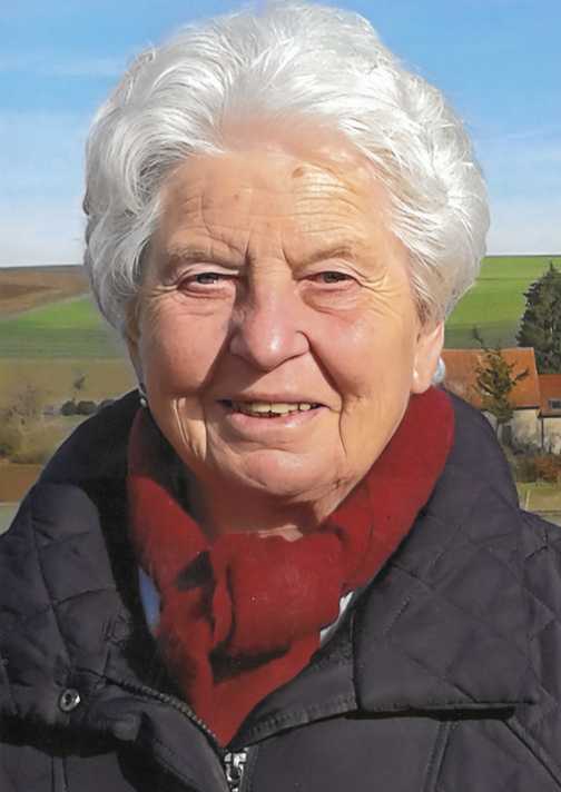 Portrait einer &auml;lteren Frau mit grauen Haaren, die l&auml;chelt und einen roten Schal tr&auml;gt, vor einer l&auml;ndlichen Landschaft.