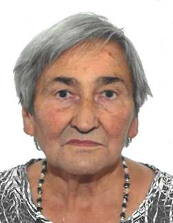 Portr&auml;t einer &auml;lteren Frau mit grauen Haaren und einem bunten Oberteil, vor hellem Hintergrund. Sie tr&auml;gt eine Halskette.