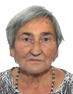 Portr&auml;t einer &auml;lteren Frau mit grauen Haaren und einem bunten Oberteil, vor hellem Hintergrund. Sie tr&auml;gt eine Halskette.