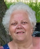 Porträt einer lächelnden älteren Frau mit lockigem, grauem Haar vor unscharfem, grünem Hintergrund.