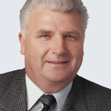 Porträt eines Mannes mit kurzem, grauem Haar und einem schwarzen Anzug, der freundlich in die Kamera lächelt.