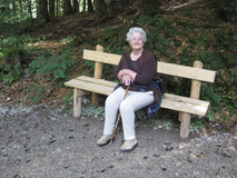 Ältere Frau sitzt auf einer Holzbank im Wald und hält einen Stock, umgeben von Blättern und grünen Pflanzen.