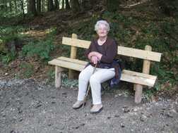 &Auml;ltere Frau sitzt auf einer Holzbank im Wald und h&auml;lt einen Stock, umgeben von Bl&auml;ttern und gr&uuml;nen Pflanzen.