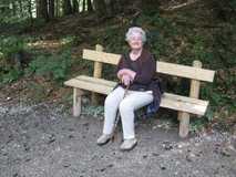 &Auml;ltere Frau sitzt auf einer Holzbank im Wald und h&auml;lt einen Stock, umgeben von Bl&auml;ttern und gr&uuml;nen Pflanzen.