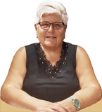 Lächelnde Frau mit kurzen, weißen Haaren und Brille, trägt ein schwarzes Oberteil und sitzt an einem Tisch.