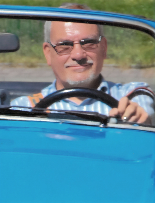 Mann mit Brille steuert ein Oldtimer-Auto, l&auml;chelnd hinter dem Lenkrad, auf einer sonnigen Stra&szlig;e.