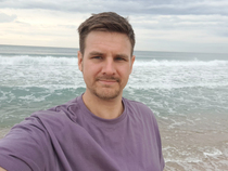Mann mit lila T-Shirt steht am Strand, Wellen schlagen sanft ans Ufer, bewölkter Himmel im Hintergrund.