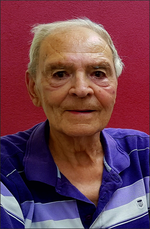 Portr&auml;t eines &auml;lteren Mannes mit grauen Haaren, der in einem lila gestreiften Hemd vor einer roten Wand sitzt.