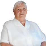 Ältere Frau mit kurzer, blonder Haarfrisur, trägt ein weißes Hemd und eine Perlenkette, steht vor neutralem Hintergrund.