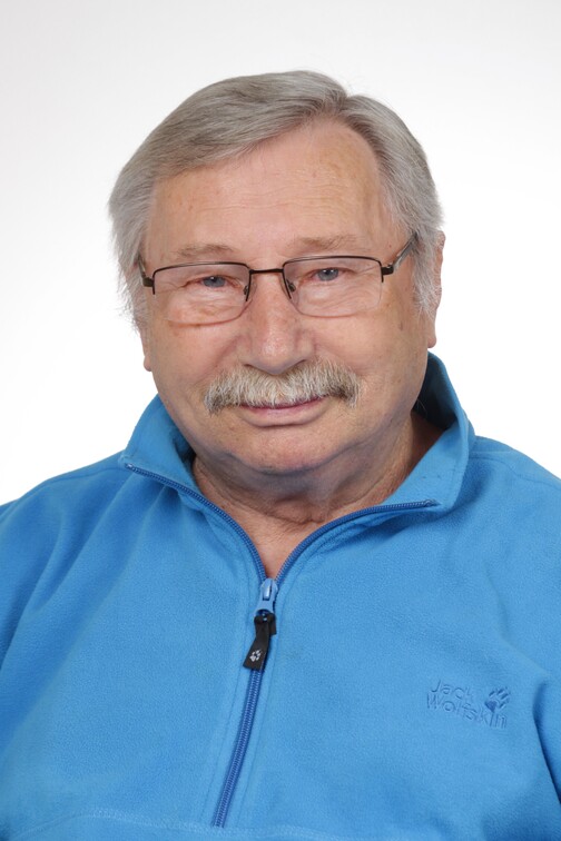 Portr&auml;t eines &auml;lteren Mannes mit Brille, grauen Haaren und Schnurrbart, der einen blauen Fleece-Pullover tr&auml;gt.