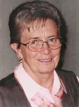 L&auml;chelnde Frau mit Brille, kurzem Haar und Ohrringen, in rosa Bluse und Weste vor neutralem Hintergrund.