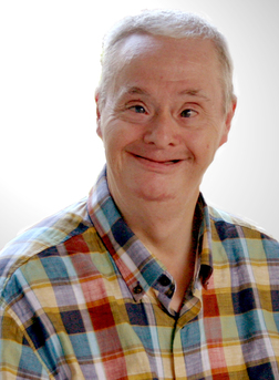 Mann mit Down-Syndrom l&auml;chelt vor einem hellen Hintergrund, tr&auml;gt ein kariertes Hemd in bunten Farben.