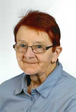 Portr&auml;t einer &auml;lteren Frau mit rotem Haar, Brille und blauer Bluse vor einem neutralen Hintergrund.