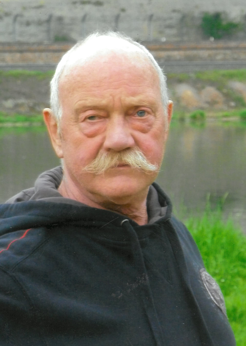 Portr&auml;t eines &auml;lteren Mannes mit grauem Schnurrbart und Kapuzenjacke, stehend vor einem Gew&auml;sser mit gr&uuml;ner Ufervegetation.