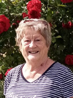 L&auml;chelnde &auml;ltere Frau in gestreiftem Shirt vor bl&uuml;henden roten Rosen im Hintergrund.