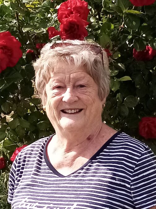 L&auml;chelnde &auml;ltere Frau in gestreiftem Shirt vor bl&uuml;henden roten Rosen im Hintergrund.