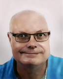 Nahaufnahme eines lächelnden Mannes mit Brille, kurzem, lichtem Haar und blauem Oberteil vor einem hellen Hintergrund.