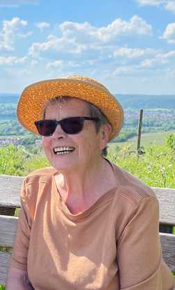 L&auml;chelnde Frau mit Sonnenhut und Sonnenbrille sitzt auf einer Holzbank vor einer gr&uuml;nen Landschaft und blauem Himmel.