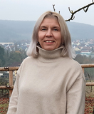 Frau mit blonden Haaren in einem beigefarbenen Rollkragenpullover steht vor einer malerischen Landschaft.
