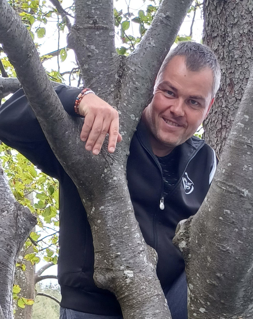 Mann lehnt l&auml;ssig an einem Baumstamm und schaut l&auml;chelnd aus dem Ge&auml;st heraus, umgeben von gr&uuml;nen Bl&auml;ttern.