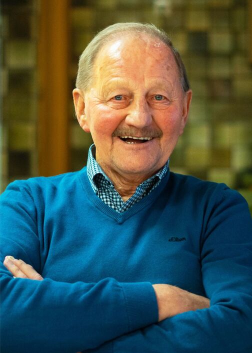 L&auml;chelnder &auml;lterer Mann mit blauer Pullover und kariertem Hemd, vor verschwommenem Hintergrund.